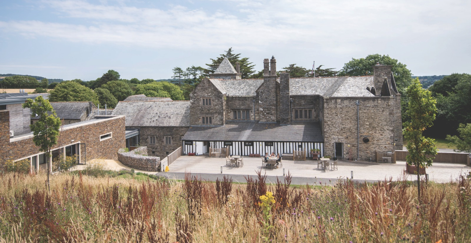 Historic stone manor house with slate roofs, tall chimneys and a black-and-white timbered ground floor overlooking a paved terrace with tables and chairs. Wild grasses and flowers fill the foreground, with leafy trees surrounding the buildings beneath a lightly clouded sky